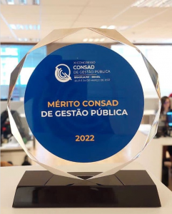 A foto mostra um troféu com base preta, fundo transparente e, no centro, um selo em azul com letras amarelas e brancas, em que se lê "Mérito CONSAD de Gestão Pública 2022". Acima, em branco, dentro do selo, vemos o logo da CONSAD