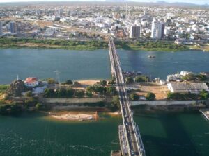 A foto mostra uma imagem aérea de uma ponte que liga duas cidades: Petrolina (PE) e Juazeiro (BA). Embaixo da ponte, vê-se um rio de cor verde