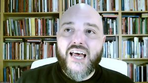 A imagem mostra um homem careca e barbudo sentado, falando. Atrás dele, vemos uma estante cheia de livros
