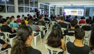 A imagem mostra várias pessoas sentadas em um auditório, atentas à uma apresentação feita por um rapaz usando uma camisa lilás